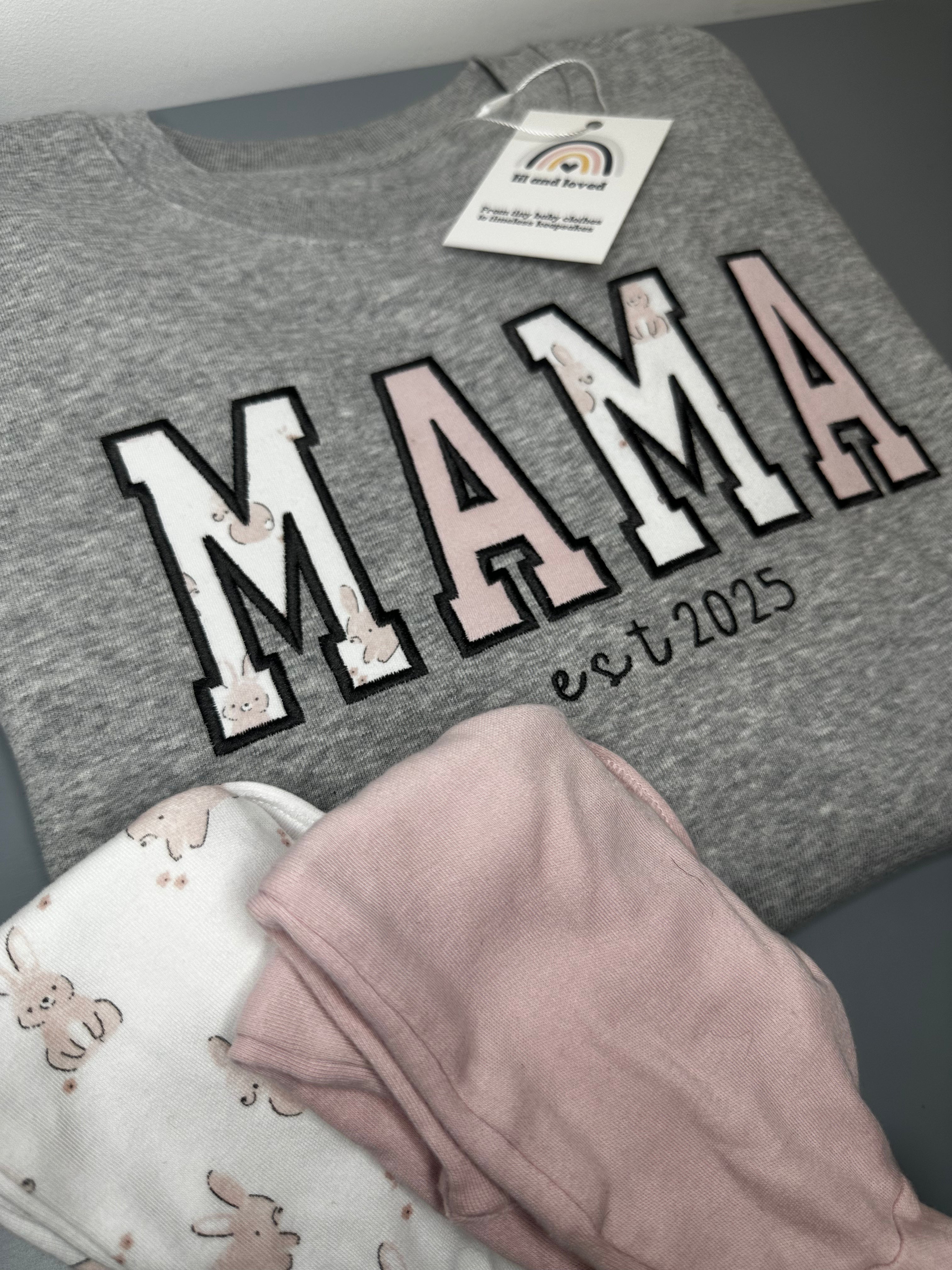 Gray sweatshirt with 'MAMA' text and baby rompers on a gray surface