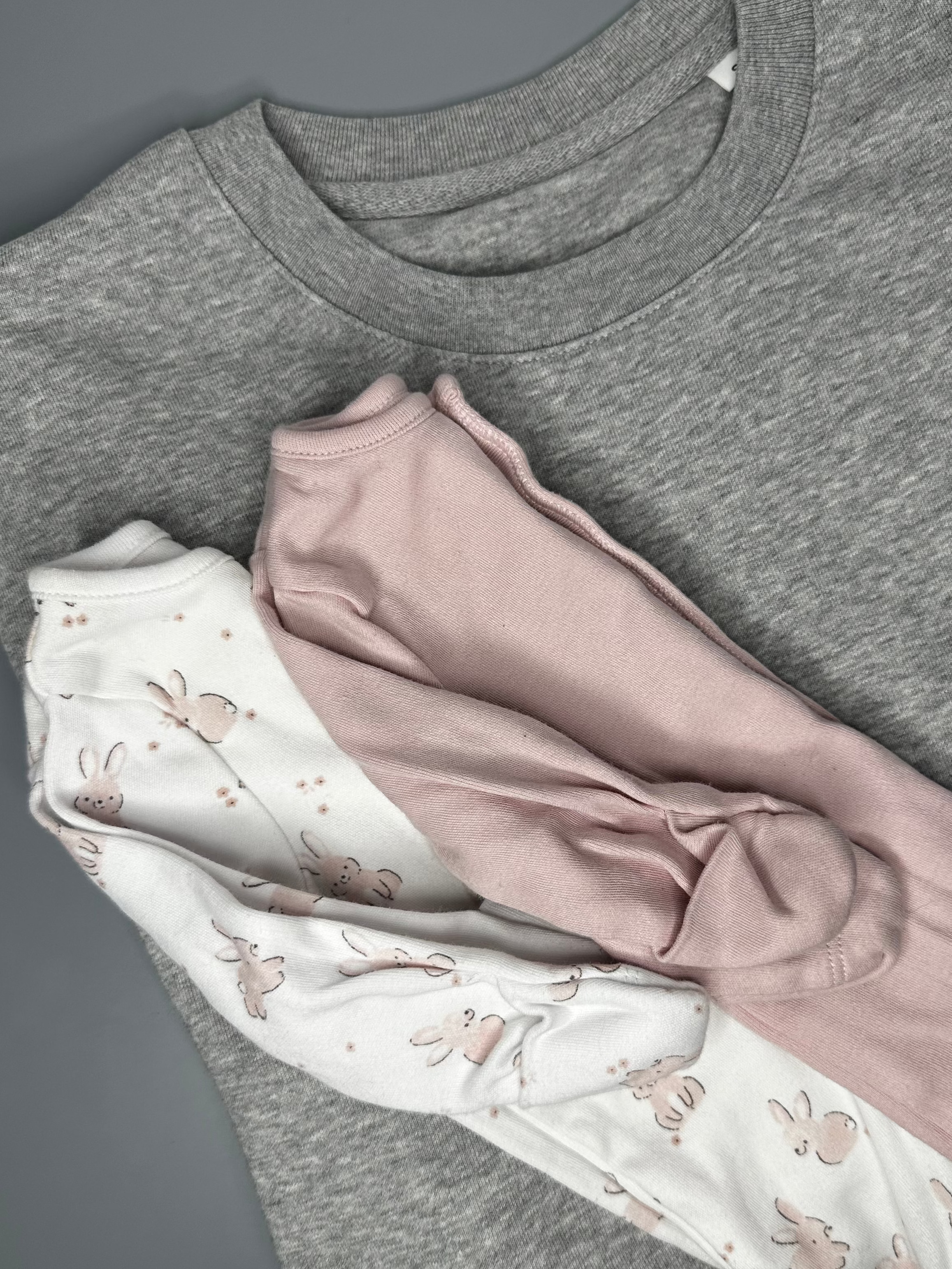 A folded heather gray long-sleeve top layered under a light pink long-sleeve baby romper and a white long-sleeve baby romper with a pink bunny print, arranged on a dark gray surface