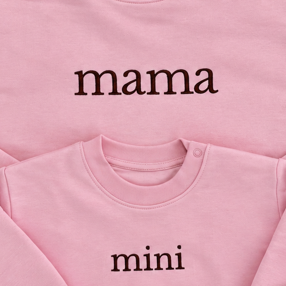 Two pink crewneck sweatshirts laid flat, one larger with the word 'mama' and one smaller with the word 'mini' in dark brown lowercase serif text.